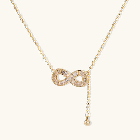 Gold infinity pendant with baguette crystals on adjustable chain against cream background.